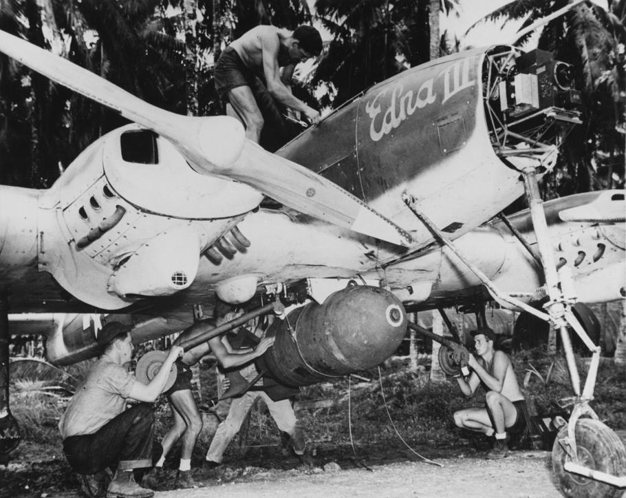 A black and white photo shows a crew of four men—two of them shirtless—working on a propellered airplane bearing the name "Edna III." The plane is parked in a tropical jungle.
