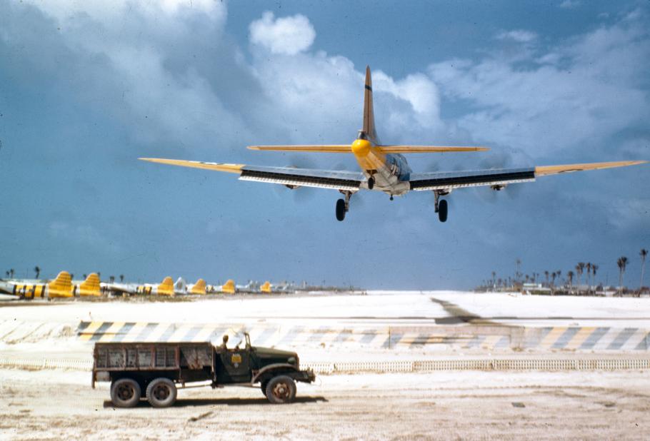 A World War II-era bomber, painted yellow, lands on a tarmac in the middle of empty desert, with an army vehicle parked in the foreground. Several other yellow-painted aircraft can be seen sitting on the tarmac in the background.