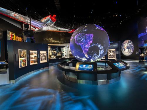 A view of the One World Connected gallery. There is a large glowing globe visible at the center of the room, images around it, and a silver and red plane hanging overhead. 