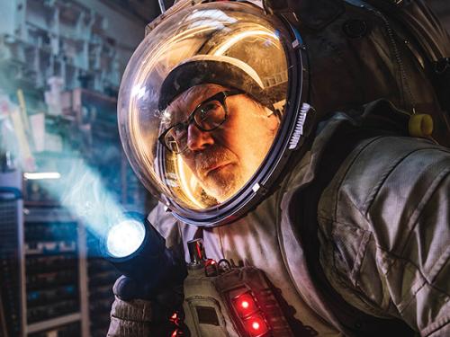 Adam Savage poses heroically in his spacesuit, a giant model of a hammerhead shark is suspended overhead. His face, wearing glasses, is illuminated behind the transparent plastic bubble that forms most of the helmet.