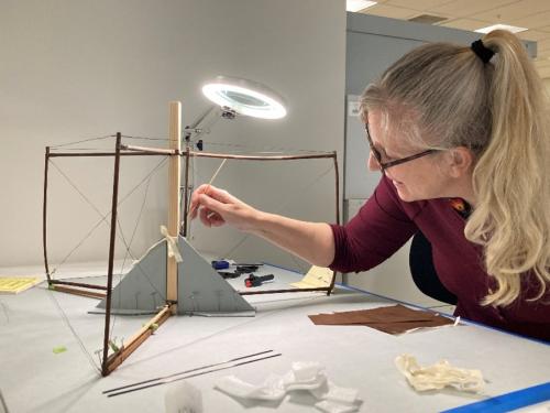 A person is engaged in assembling a kite structure inside a room, focusing intently on adjusting a component of the structure.