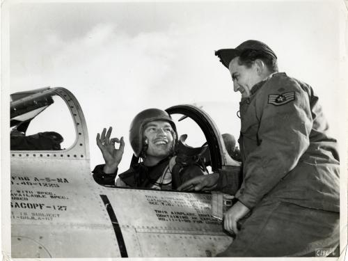 Man in a helmet sits in an aircraft cockpit giving the okay sign with his right hand to a man in a cap and flight suit on his left
