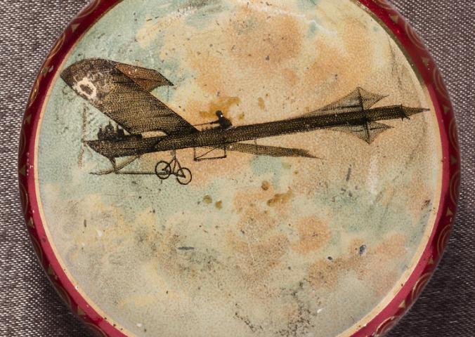 Circular metal box with monoplane and pilot in a cloudy sky painted on the lid. Image surrounded by narrow gold border. Sides and bottom metallic red.