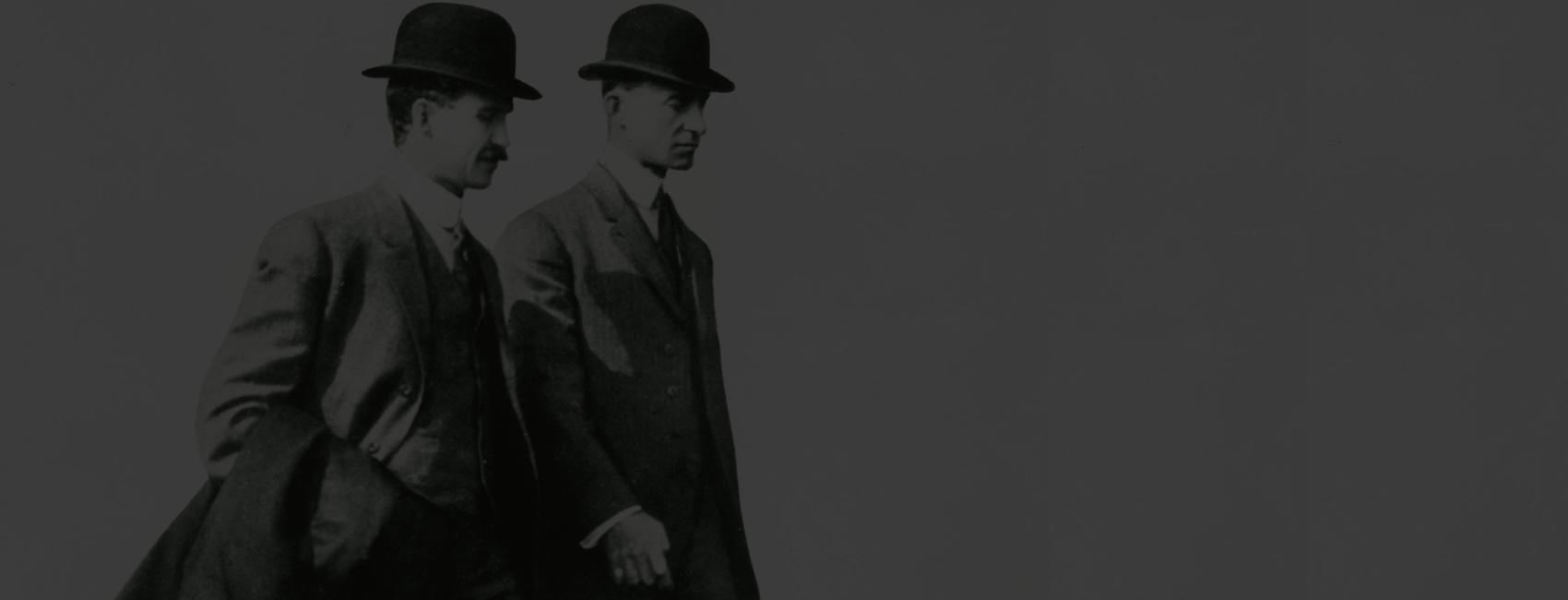 Orville and Wilbur Wright, seen from the side, walk next to each other, against a blank grey background.