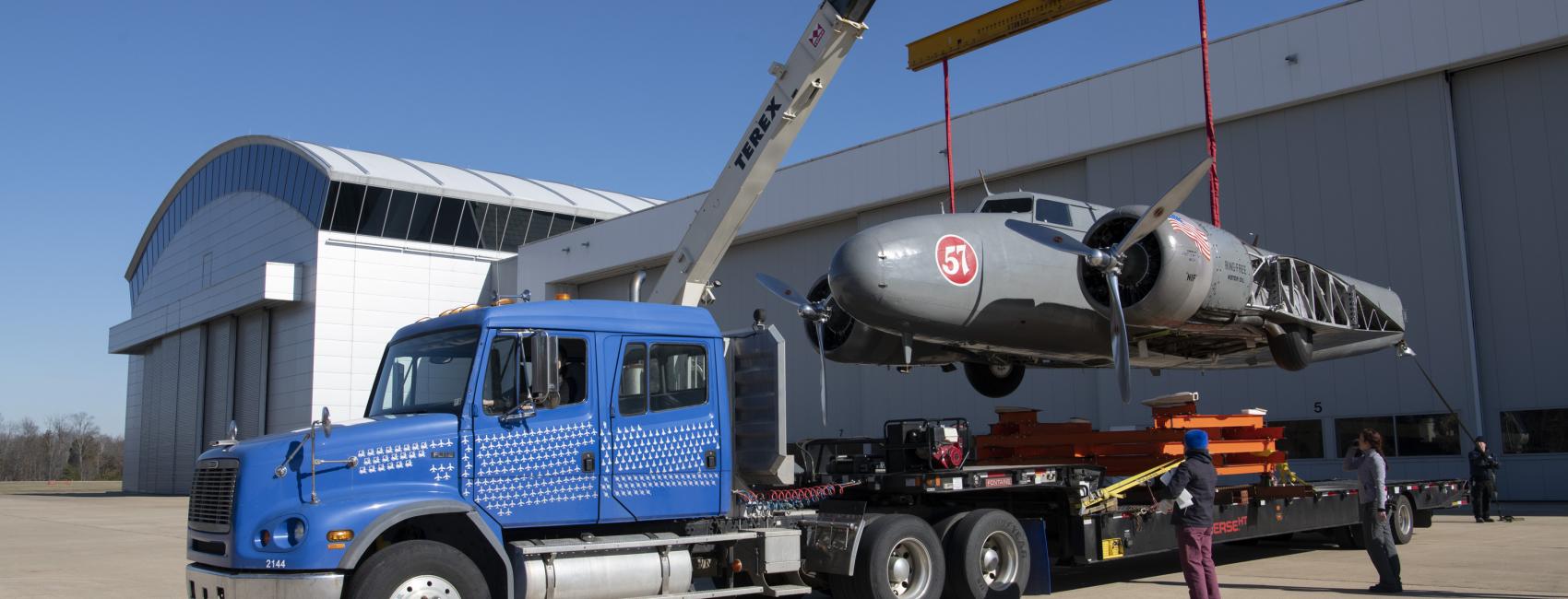 A crane lowers a large airplane body onto the bed of a flatbed truck. 
