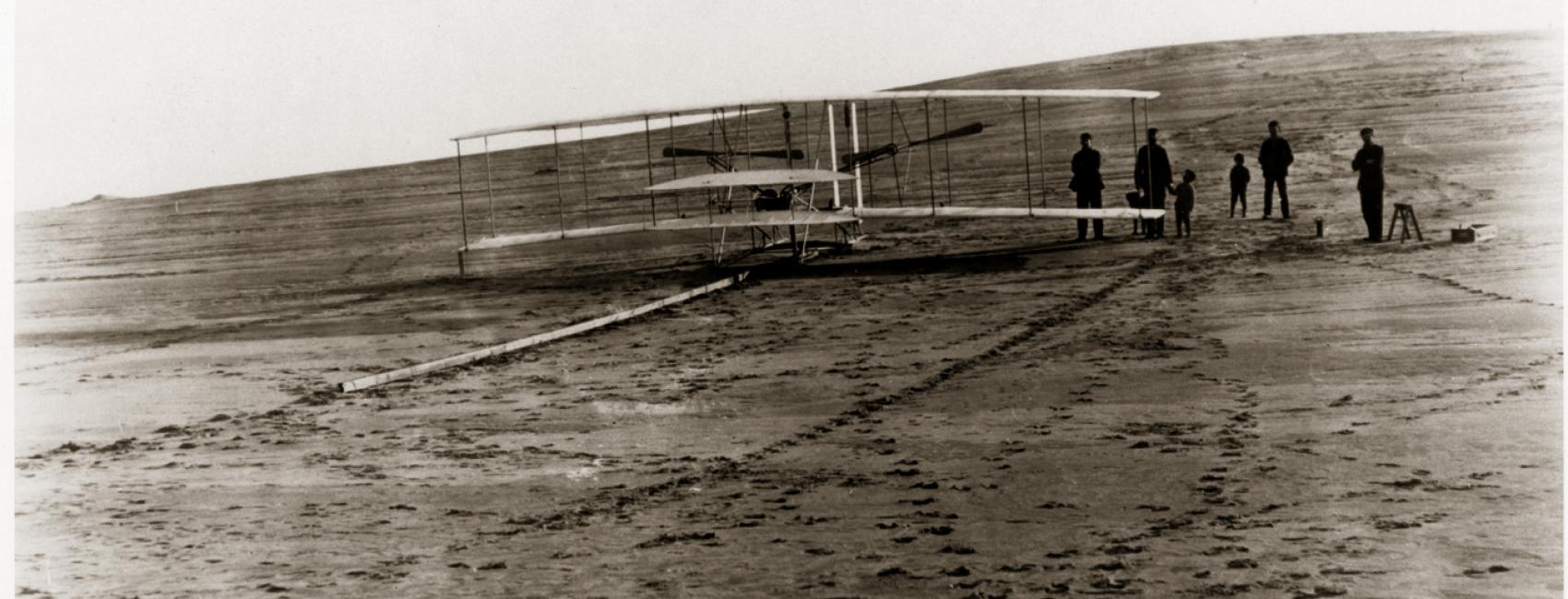 A biplane sits on a long wooden rail on the ground. A group of people stand behind it.