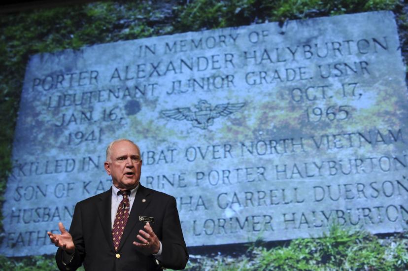 Senior man in suit and tie gesturing with his hands. A slide with engraved text is behind him..
